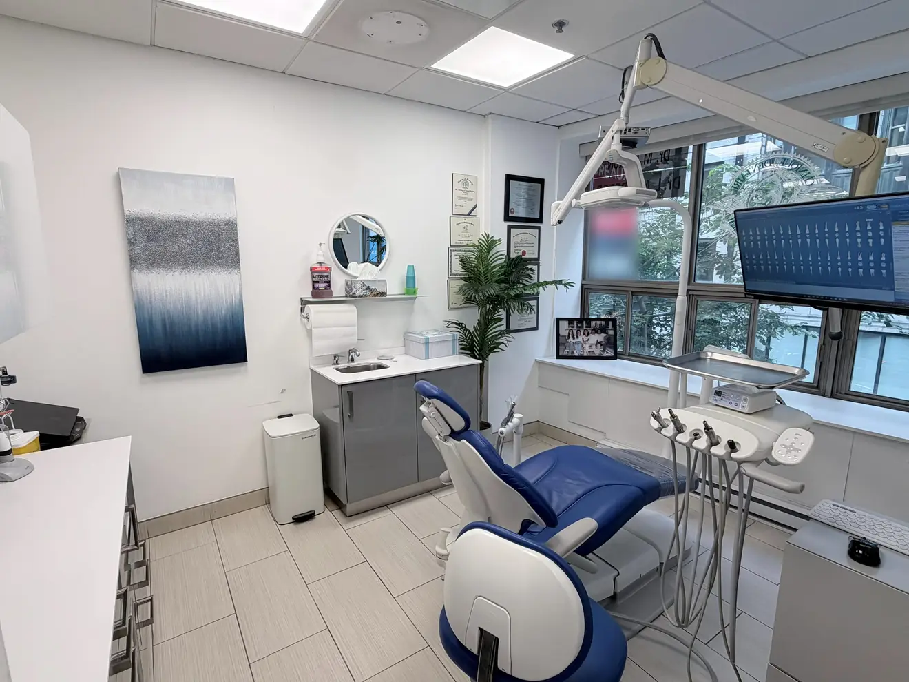 A bright treatment room: a blue dental chair, a screen above it and large windows onto the street